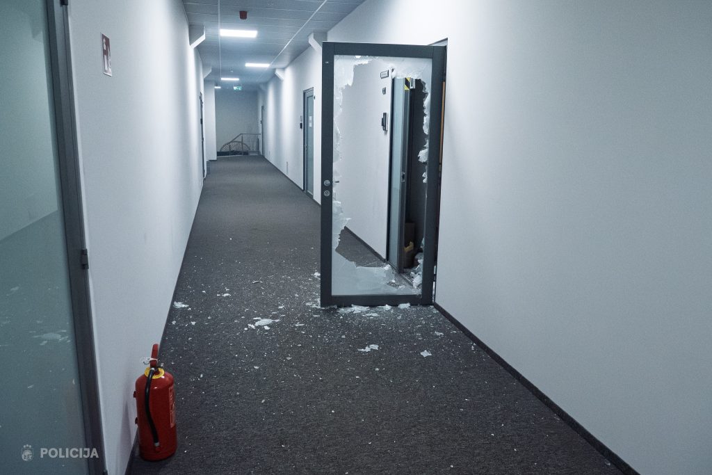 Tukšs biroja koridors ar tumšu paklāju un atvērtām durvīm, kurām izsists stikls un stikla lauskas uz grīdas, norādot uz policijas ielaušanos.