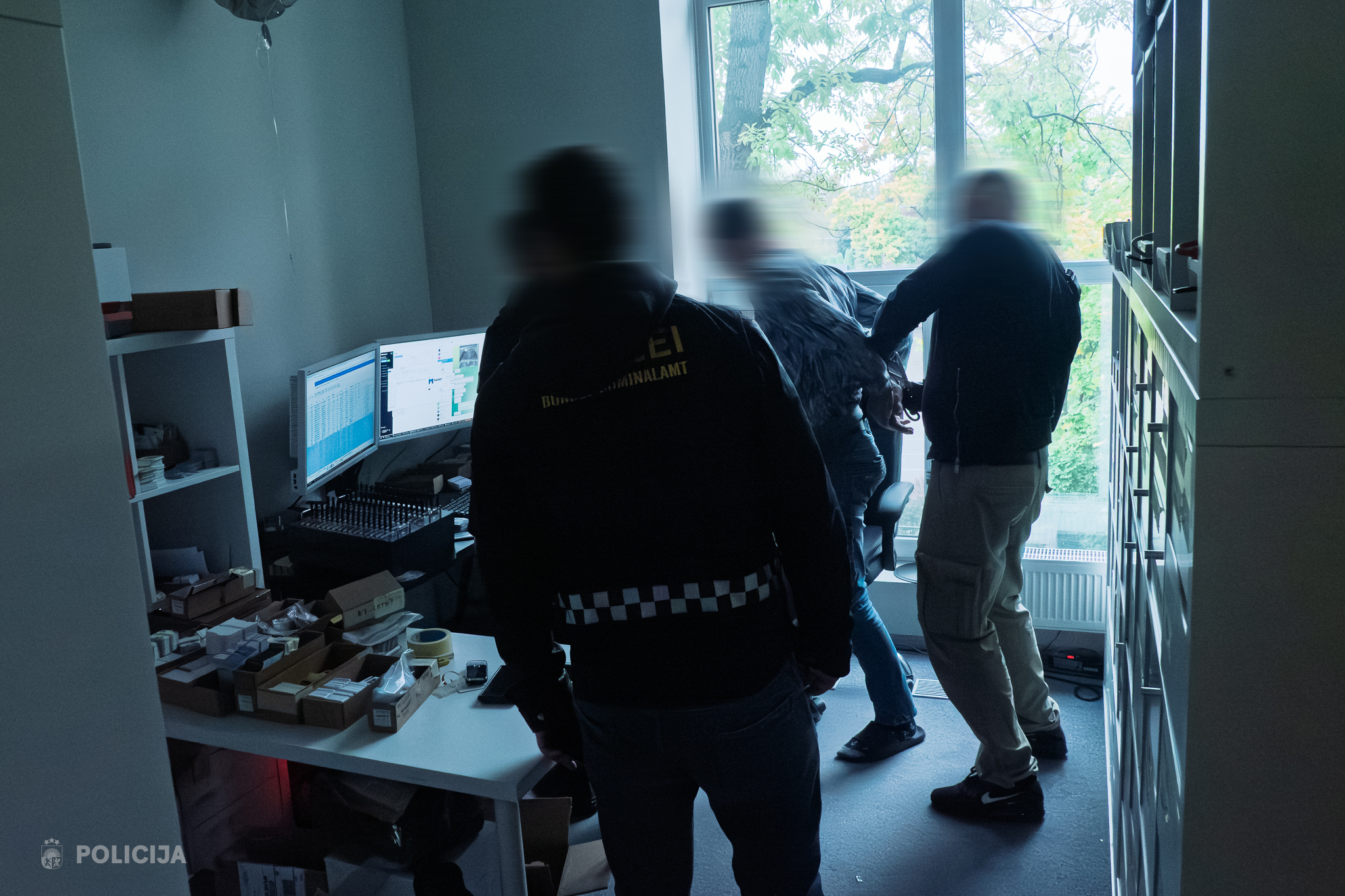 Valsts policijas darbinieki (aizmigloti) veic aizturēšanu gaišā telpā pie loga, kur fona gaismas rada kontrasta efektu.