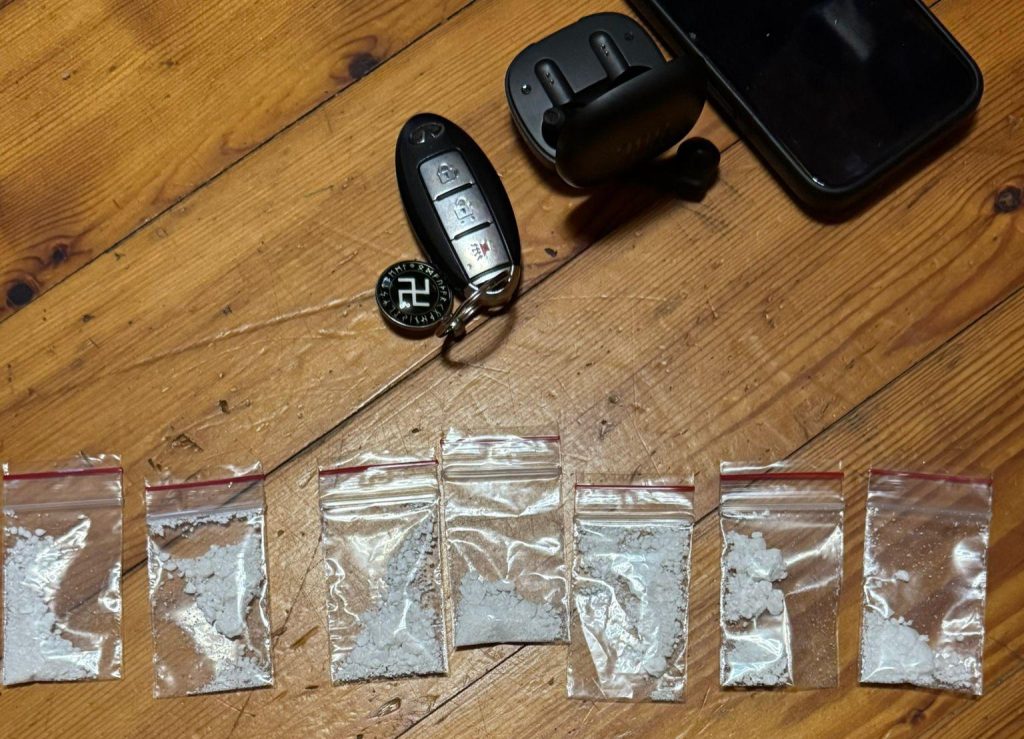 Septiņi caurspīdīgi maisiņi ar baltu pulveri (kokaīnu/alfa-PVP), novietoti uz koka galda blakus automašīnas atslēgai un austiņām.