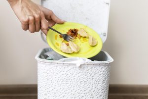 Persona ar dakšu izmet ēdiena atliekas (pelmeņus un dārzeņus) no šķīvja atkritumu tvertnē.