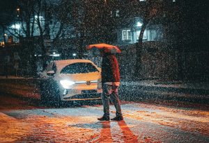 Gājējs ar lietussargu sniegputenī naktī pie balta automobiļa ar ieslēgtām gaismām pilsētā.