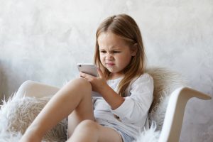 Maza meitene ar sarauktām uzacīm un neapmierinātu sejas izteiksmi sēž pūkainā krēslā un skatās mobilajā telefonā.