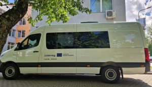 Balts mobilais klientu apkalpošanas mikroautobuss ar "Interreg" un ES simboliku, kas stāv pie ēkas Līvānos.