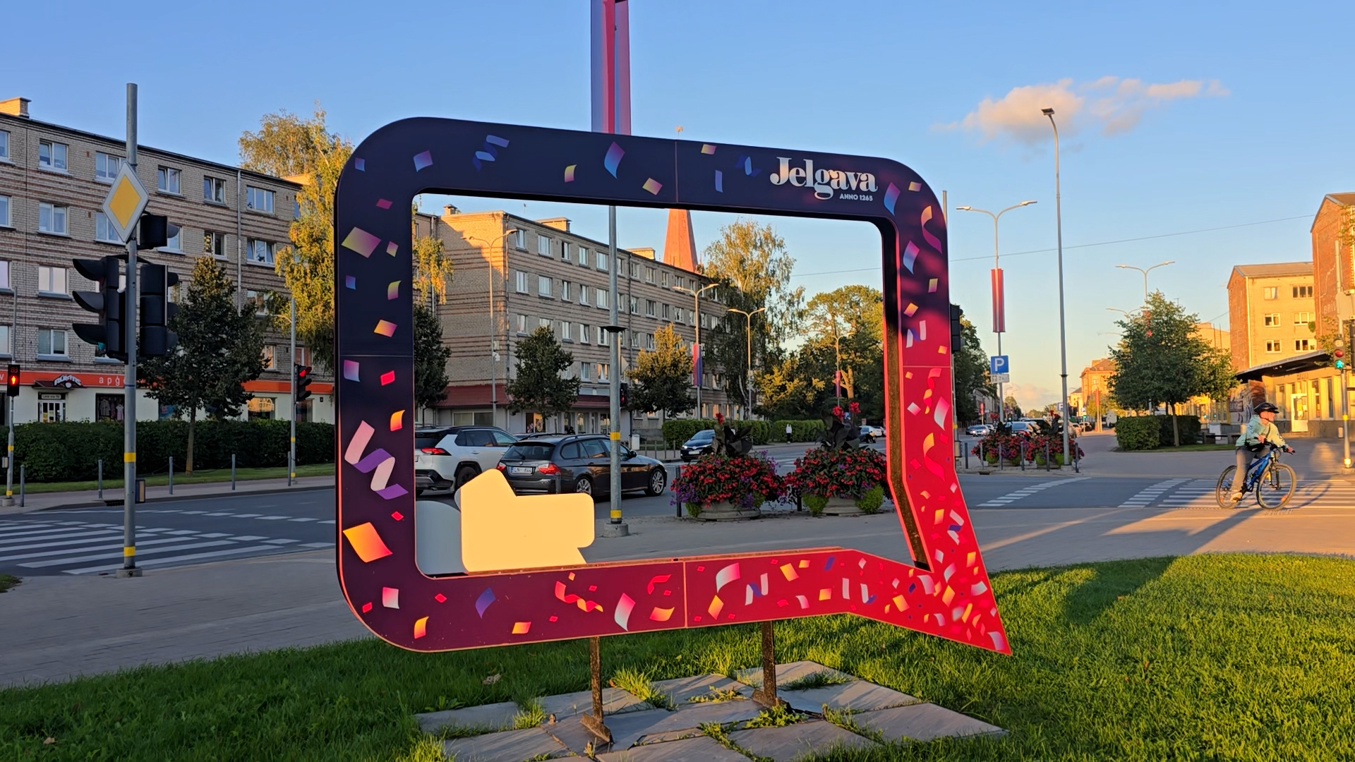 Liela, krāsaina Jelgavas pilsētas fotokadra instalācija ielas malā ar daudzdzīvokļu māju fonā.