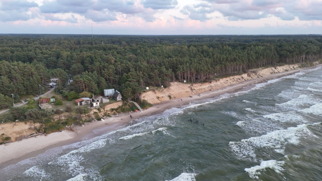 Panorāmas skats no augšas uz Jūrkalnes stāvkrastu, priežu mežu un Baltijas jūru ar nelielām ēkām krastā.