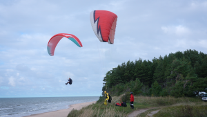 Divi paraplāni lido virs Jūrkalnes stāvkrasta, bet divi piloti stāv uz pakalna, gatavojoties lidojumam vai vērojot.