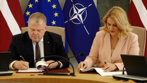 Evika Siliņa un Gints Kaminskis sēž pie galda, katrs parakstot dokumentu uz sarkanām mapēm.