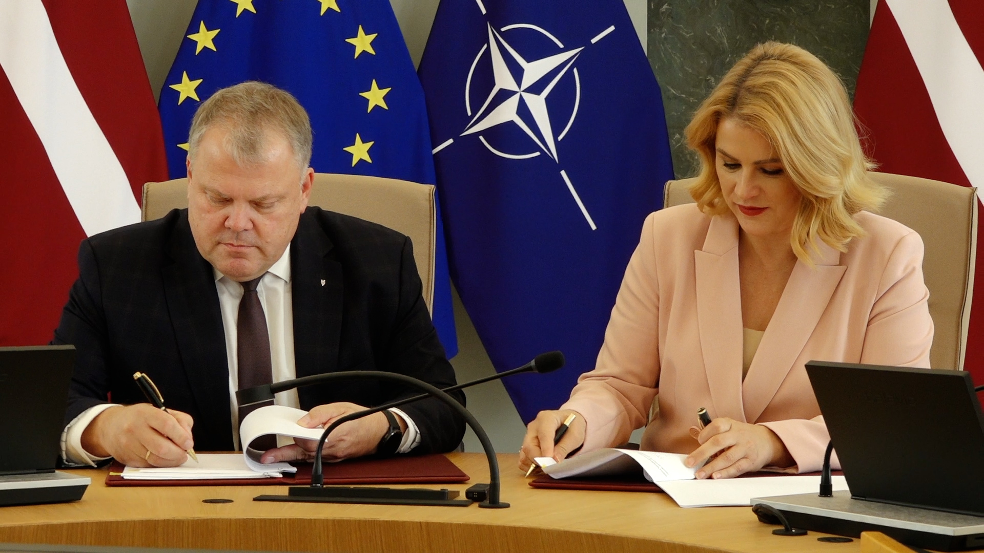 Evika Siliņa un Gints Kaminskis sēž pie galda, katrs parakstot dokumentu uz sarkanām mapēm.