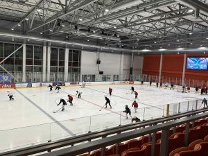 Skats uz hokeja laukumu, kur sieviešu komanda trenējas. Fonā redzams liels video ekrāns ar sporta saturu.