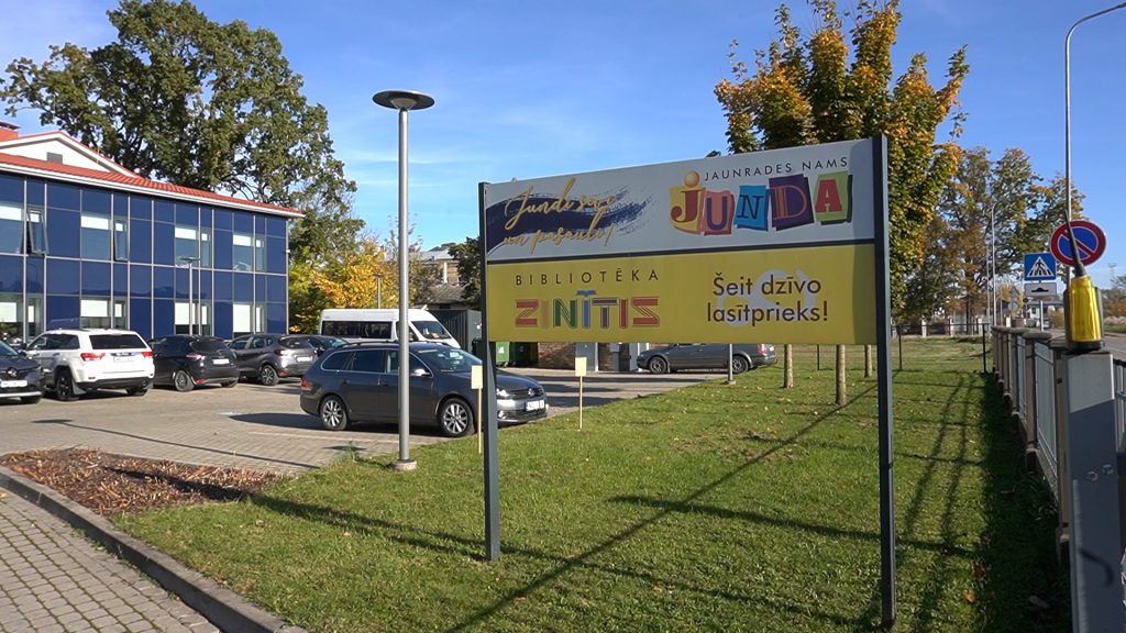 Jaunrades nama "Junda" izkārtne ar bibliotēkas "Zinītis" nosaukumu. Fonā redzama moderna stiklotas ēkas fasāde un autostāvvieta.