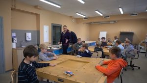 Zēni pie koka galdiem, pieliekušies pie inerces auto modeļiem un citām detaļām Jelgavas Jaunrades nama "Junda" darbnīcā. Pie galdiem stāv skolotājs.