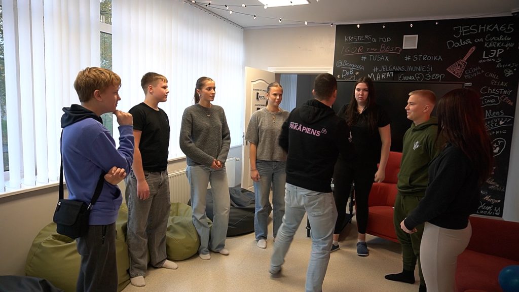 Grupa pusaudžu stāv aplī un sarunājas ar vadītāju (mugurplānā) Jauniešu iniciatīvu centrā "Pietura".
