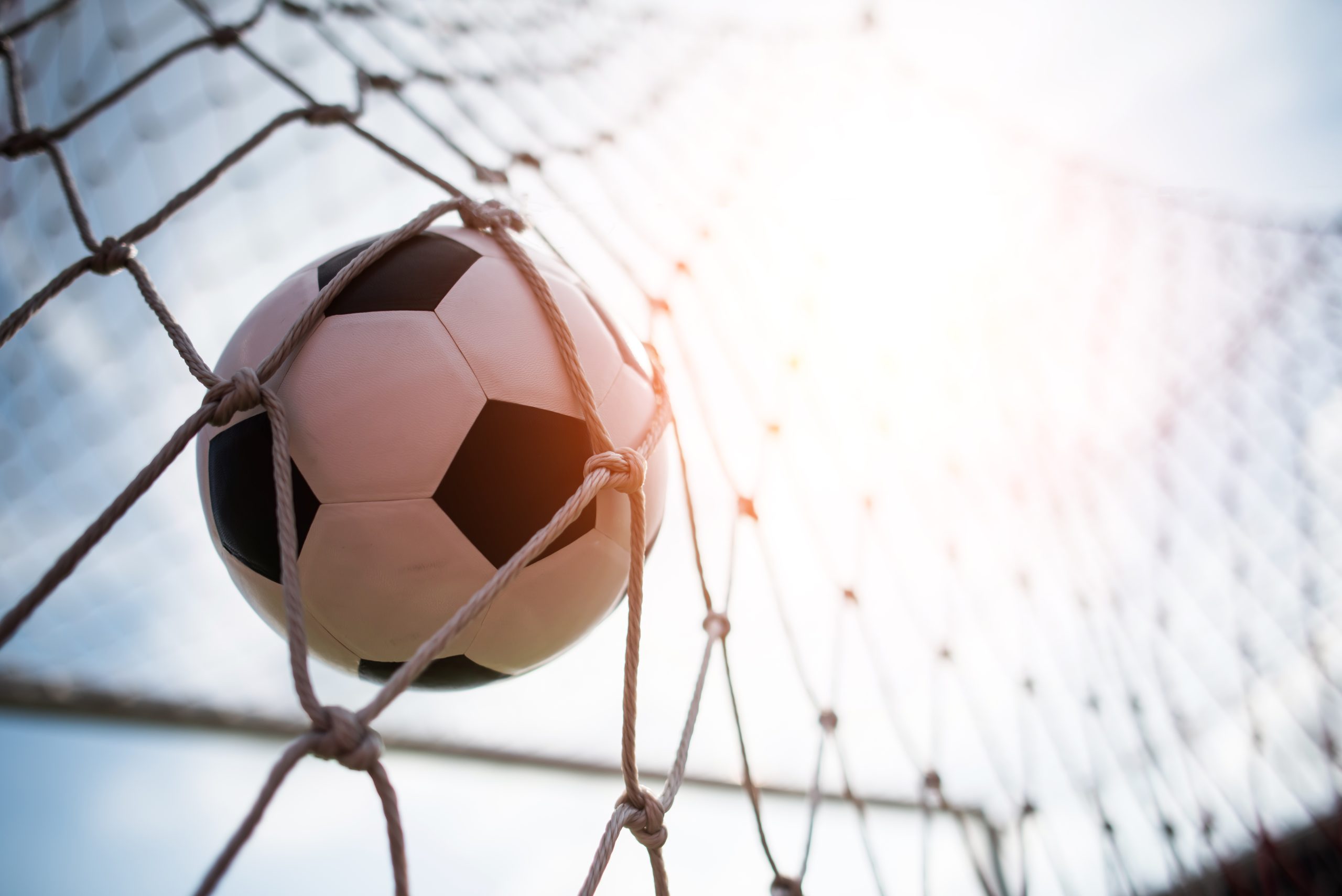 Klasiska futbola bumba melnbaltā krāsā ir ielidojusi vārtu tīklā, fonā spīd spoža saules gaisma.