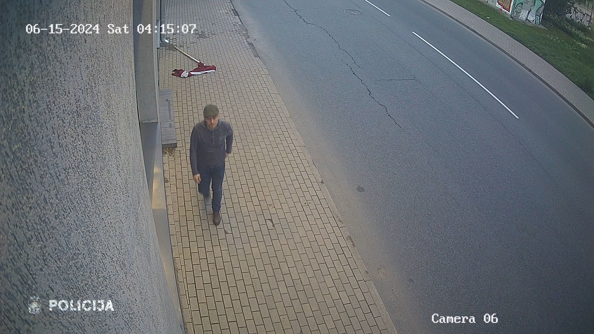 Videonovērošanas attēls, kurā redzams vīrietis, kas iet pa ietvi, bet uz zemes mētājas Latvijas karogs.