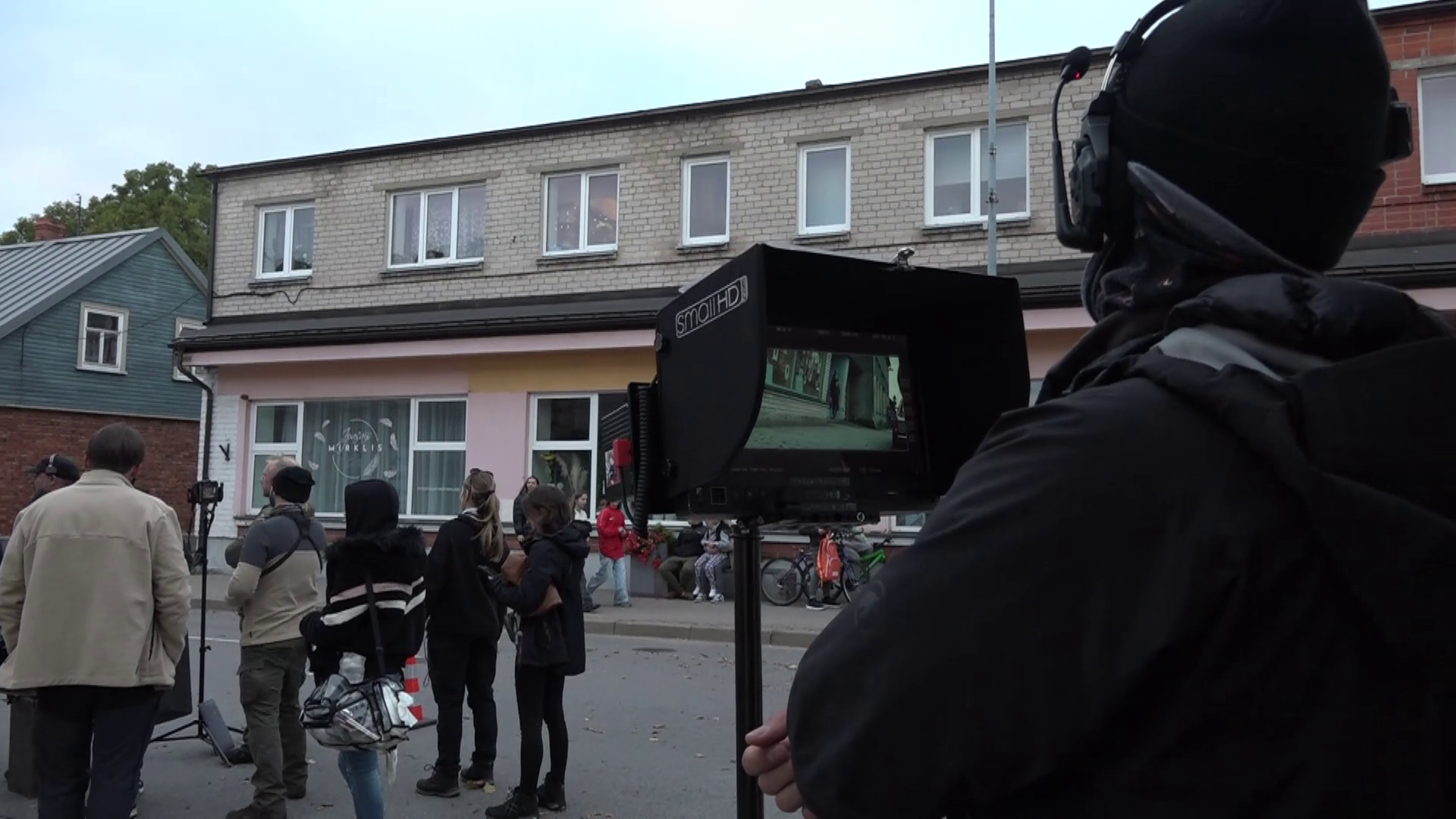 Filmēšanas komanda strādā uz ielas, viens operators vēro kadru monitorā ar ainu no filmēšanas laukuma.