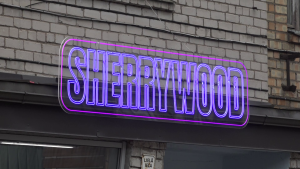 Ēkas fasāde ar spilgtu violetu neona izkārtni “SHERRIWOOD”.