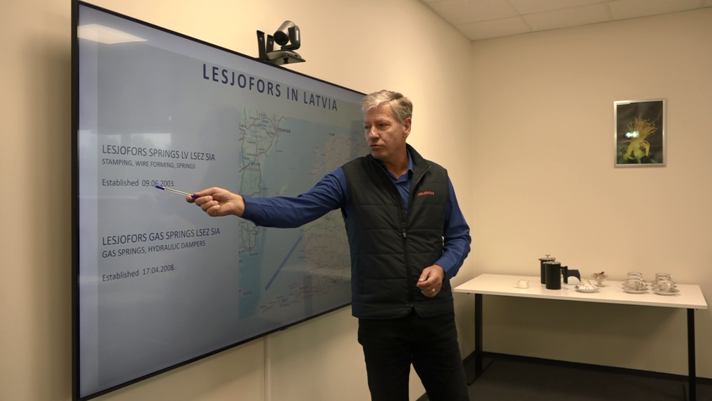 LSEZ SIA “Lesjofors Springs LV” pārstāvis rāda ar pildspalvu uz liela ekrāna, uz kura redzama uzņēmuma informācija Latvijā.
