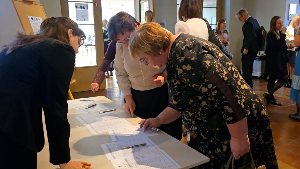 Tuvplānā vairākas sievietes noliekusies paraksta dokumentus uz reģistrācijas galda.