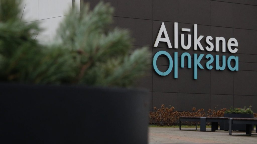 Tuvplāns uz baltu un tirkīza krāsas uzrakstu "Alūksne" uz tumšas ēkas fasādes, priekšplānā izplūdis augs.