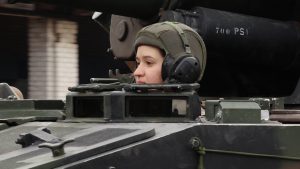 Valsts aizsardzības dienesta karavīre Glorija Sidorova ar bruņucepuri un austiņām izbāzusies no militārā transportlīdzekļa lūkas.