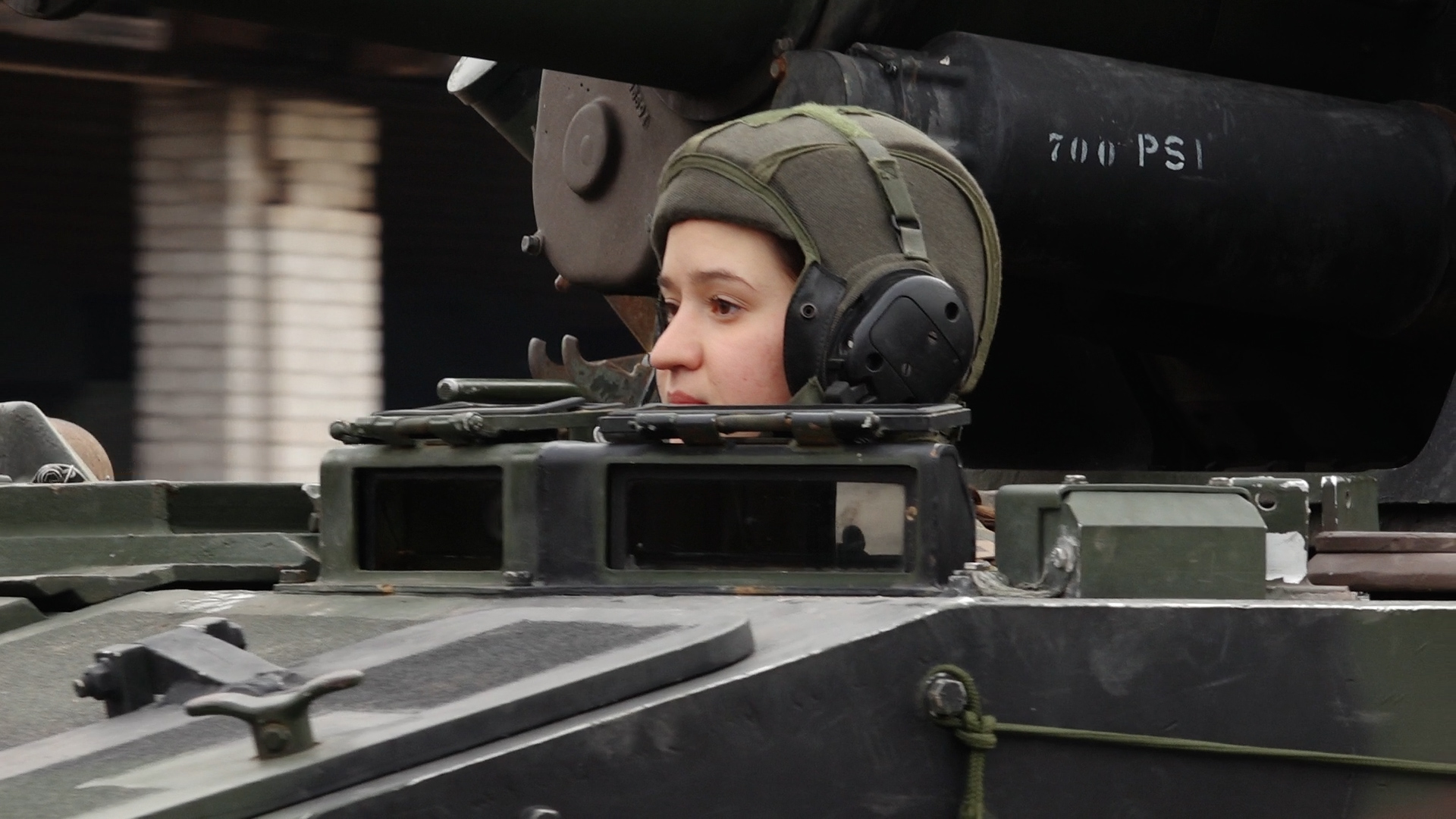 Valsts aizsardzības dienesta karavīre Glorija Sidorova ar bruņucepuri un austiņām izbāzusies no militārā transportlīdzekļa lūkas.