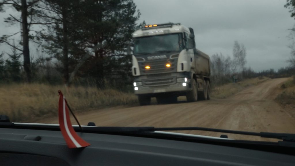 Kravas auto brauc pa grants ceļu pretī automašīnai, no kuras uzņemts attēls, ar Latvijas karoga simbolu priekšplānā.