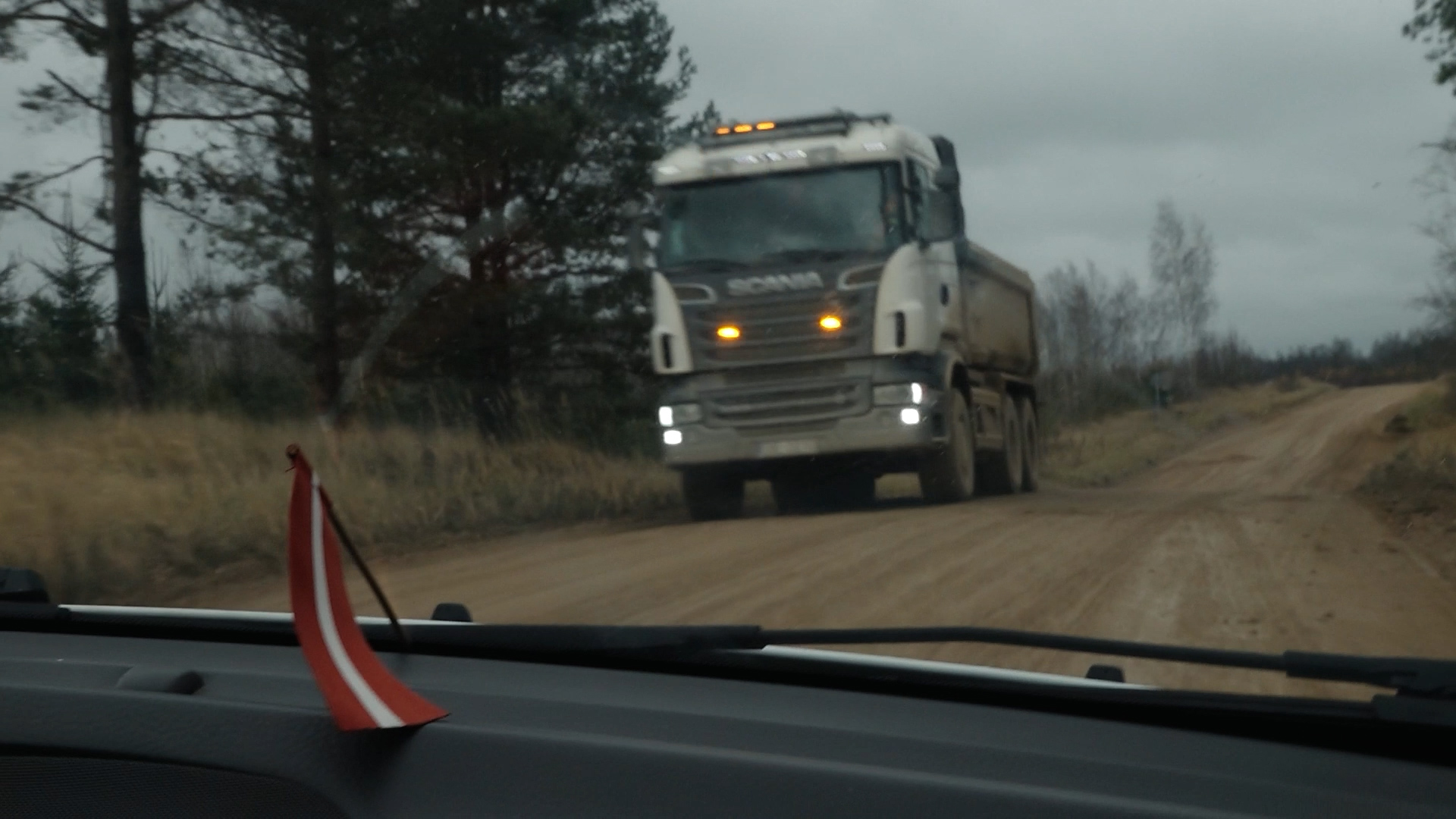 Kravas auto brauc pa grants ceļu pretī automašīnai, no kuras uzņemts attēls, ar Latvijas karoga simbolu priekšplānā.