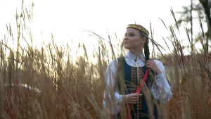 Jauna sieviete tautastērpā stāv rudens zālē ar gariem, sausiem stiebriem priekšplānā, skatoties uz augšu pret rīta gaismu.