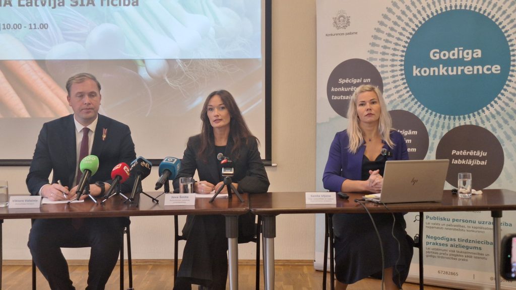Konkurences padomes pārstāvji (vīrietis un divas sievietes) sēž pie galda preses konferences laikā ar uzrakstu "Godīga konkurence".