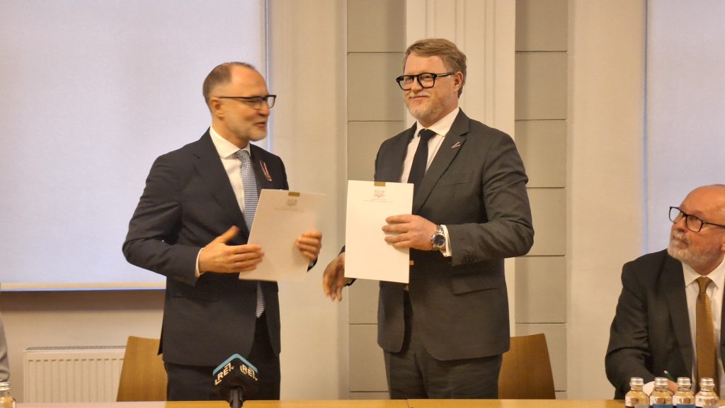 Divi vīrieši, Aizsardzības ministrs Andris Sprūds un RTU rektors Tālis Juhna, stāv kājās un sasveicinās, turot dokumentus rokās.
