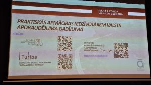 Prezentācijas slaids par praktiskajām apmācībām iedzīvotājiem valsts apdraudējuma gadījumā ar QR kodiem un organizatoru informāciju.