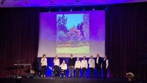 Dziedātāju grupa uz skatuves ar rudens ainavas attēlu projekcijā koncertā kultūras centrā.