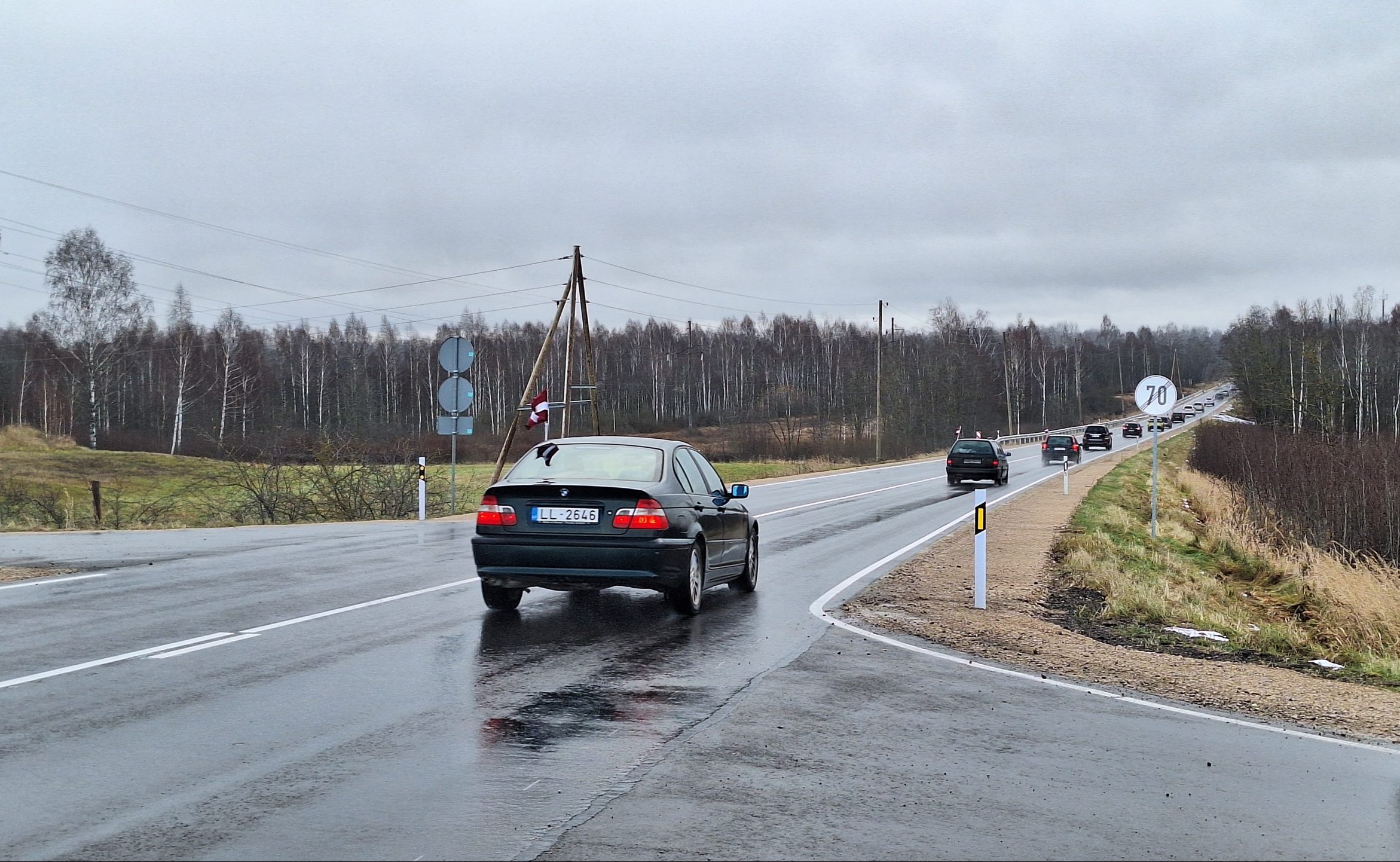 Mitrs lauku ceļš ar automašīnām un Latvijas karogu pie transporta.