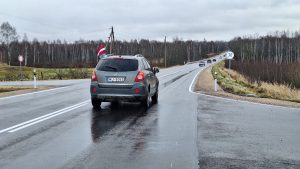Auto ar Latvijas karogu brauc pa lauku šoseju lietainā laikā.