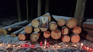 Mežā uz sakrautām baļķu rindām novietots plakāts, priekšā liesmo svecītes sniegā.