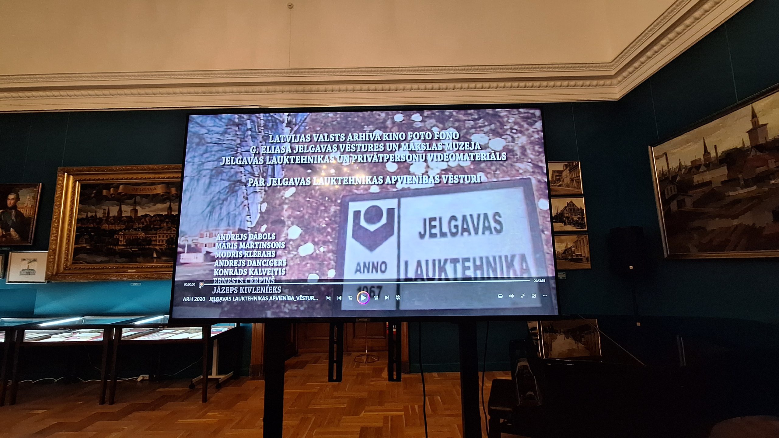 Video projekcija izstādē, kurā redzami filmas titri un uzņēmuma "Jelgavas Lauktehnika" logotips.