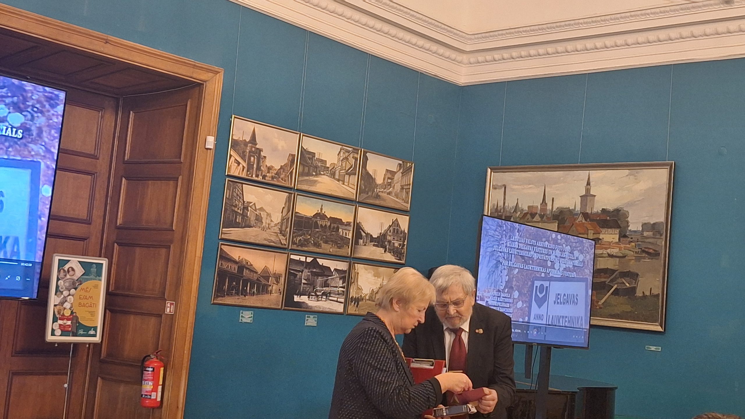 Sirms vīrietis (Jāzeps Kivlenieks) ar sarkanu kaklasaiti un sieviete stāv pie maza ekrāna gleznu izstādē.