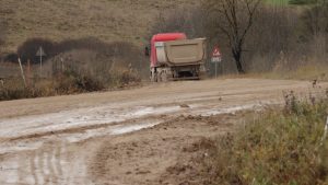 Sarkans kravas auto ar smilšu piekabi brauc pa ļoti dubļainu un izdangātu lauku ceļu, atstājot dziļas riepu sliedes.