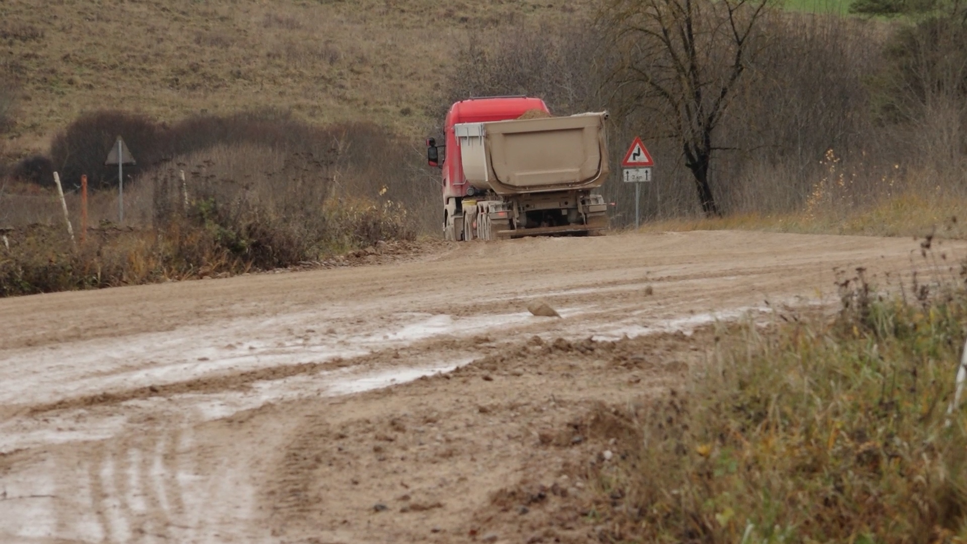 Sarkans kravas auto ar smilšu piekabi brauc pa ļoti dubļainu un izdangātu lauku ceļu, atstājot dziļas riepu sliedes.