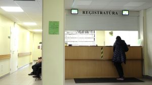 Pacients pie medicīnas iestādes reģistratūras ar uzrakstu "REĢISTRATŪRA".