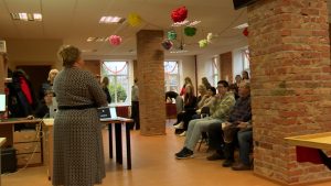 Sieviete stāv ar muguru pret kameru, runājot auditorijai, kas sēž Valkas Jauniešu biroja telpā.