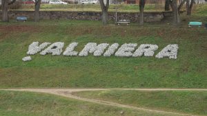 Ziedu uzraksts "VALMIERA" uz zaļas nogāzes pilsētvidē.