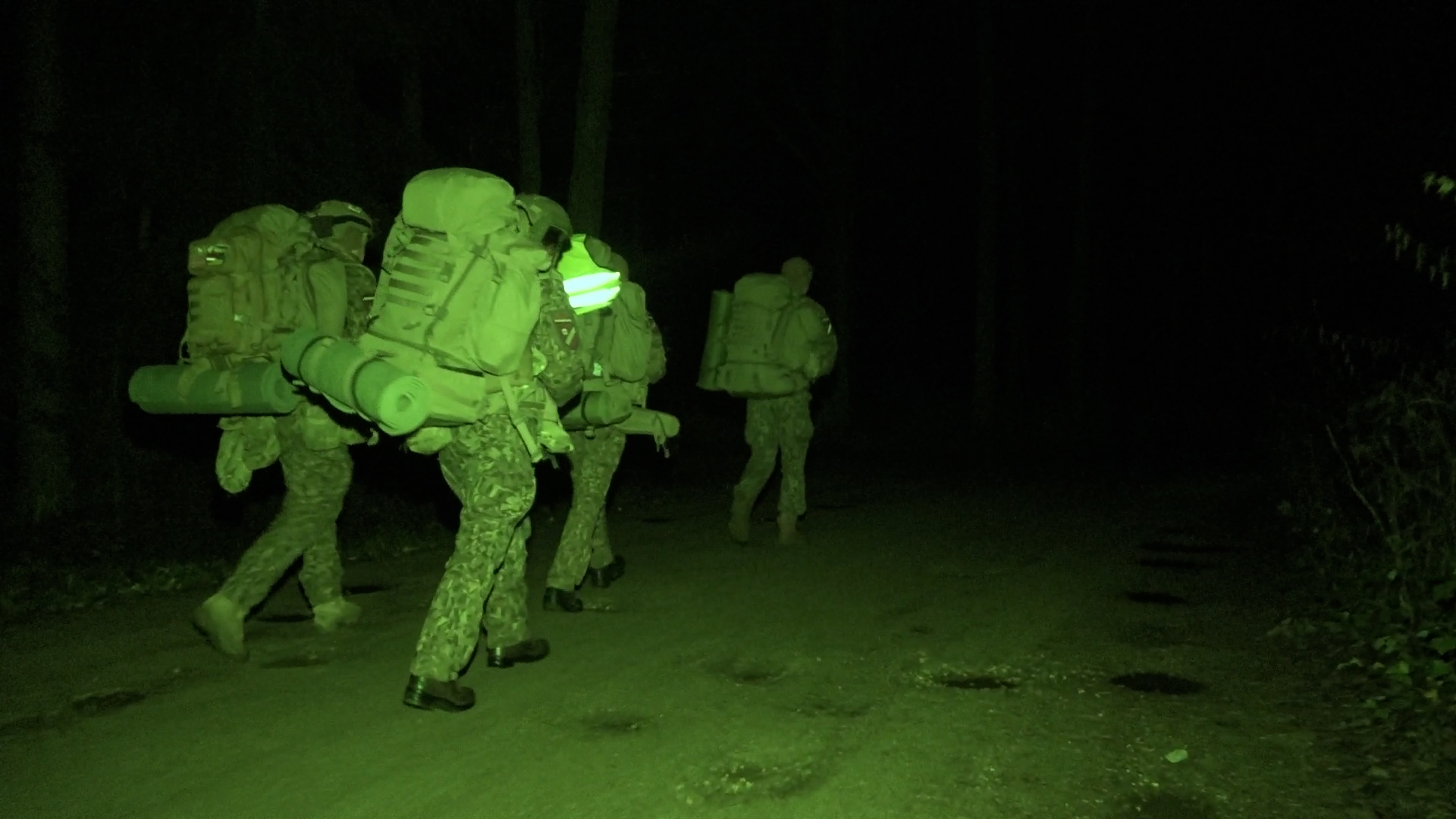 Četri karavīri militārajās uniformās ar mugursomām nakts gājienā, kadrs ar zaļu naktsredzamības filtru