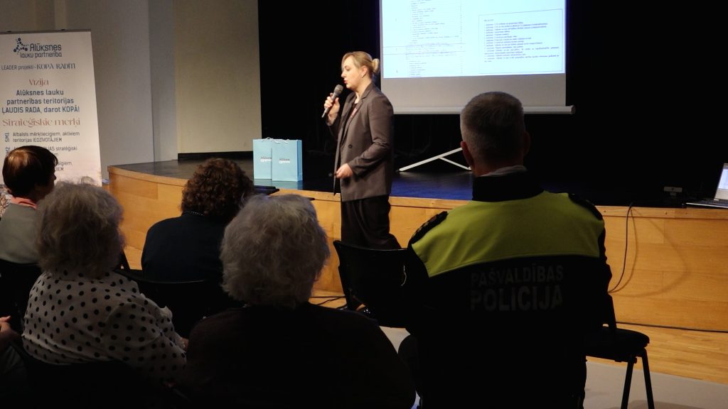 Civilās aizsardzības speciāliste Maija Sliņķe stāv uz skatuves, uzrunājot cilvēku auditoriju konferencē Alūksnē, priekšplānā Pašvaldības policists.