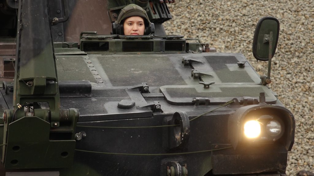 VAD karavīre Glorija Sidorova ar bruņucepuri skatās uz priekšu no haubices lūkas.