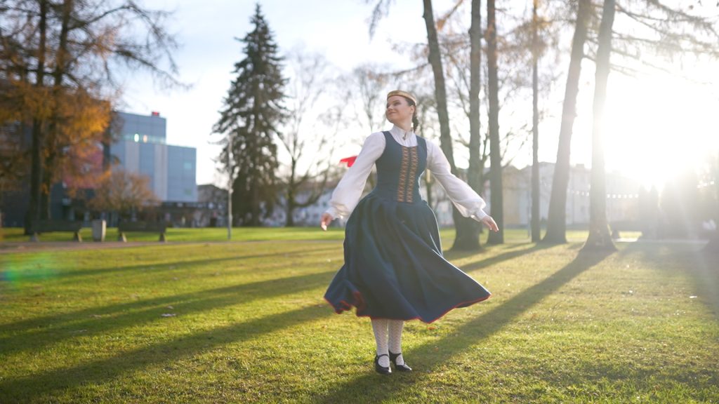 Jauna sieviete tautastērpā griežas dejas solī saulainā parkā, ap viņu veidojas garas koku ēnas.
