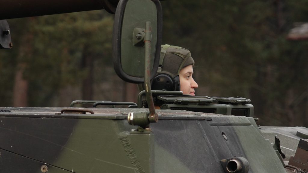 VAD karavīre Glorija Sidorova, skatoties uz priekšu no militārā transportlīdzekļa lūkas, ar zaļu meža fonu.