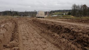 Dziļas dubļu rises un grāvji priekšplānā uz lauku ceļa ar mikroautobusu tālumā.