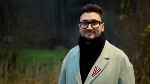 ReTV ziņu moderators Gustavs (gaiša mētelī un brillēs) smaidot pozē dabā, viņa mēteļa atlokā piestiprināta Latvijas karoga lentīte.