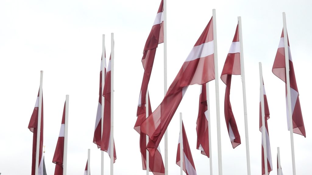Daudz Latvijas valsts karogu sarkanbaltsarkanās krāsās mastos pret gaišām debesīm.